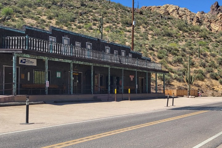 Desert building with rustic facade, cacti nearby, located alongside a road under clear blue sky.