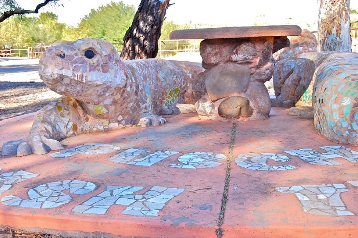 Mosaic lizard sculpture with table on back, surrounded by trees and sky.